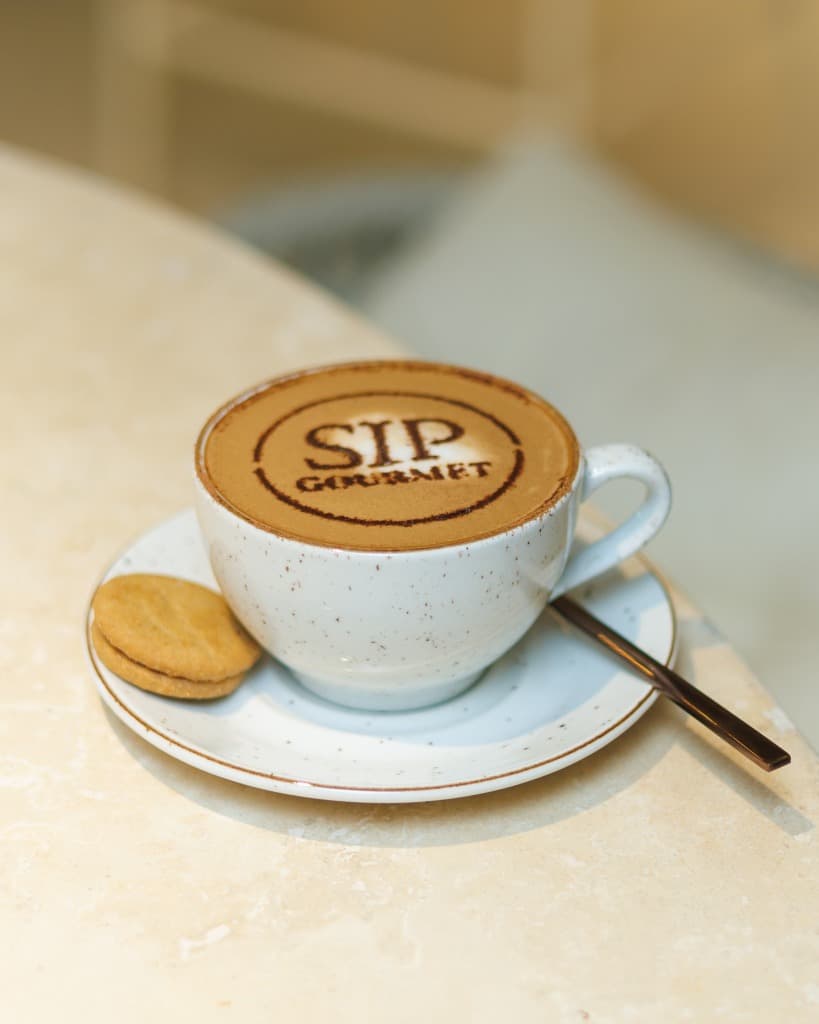 A beautifully crafted latte with 'SIP GOURMET' art, served with a delicate cookie.