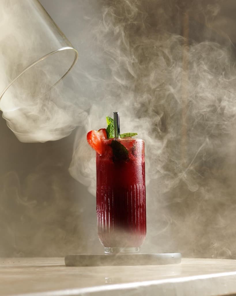 A vibrant red signature cocktail being served with a dramatic smoke effect.